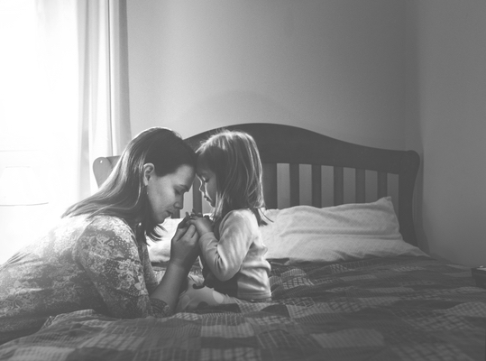 A mother and young child sitting on a bed with heads bowed and hands joined in prayer.